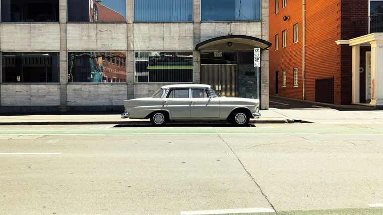 A cream Mercedes-Benz fintail sedan parked side-on against a concrete and red brick city building.
