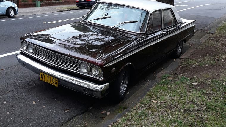Maroon Ford Fairlane sedan with a white roof parked on a tree-lined suburban street