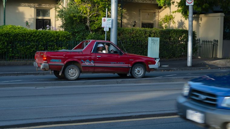 Red Subaru Brumby ute driving past ornate Victorian-era terrace houses on a tree-lined street
