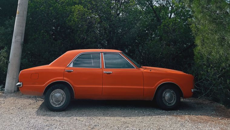 Orange 1970s-style four-door sedan parked on a gravel roadside beneath tall pine trees