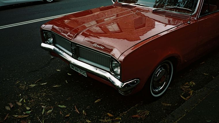 The bonnet and grille of a dark red Holden Monaro parked on a suburban street, with autumn leaves scattered across the road.