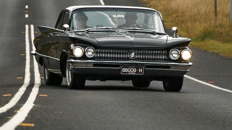 Black classic Buick sedan driving along a rural Victorian road, displaying a Victorian number plate.