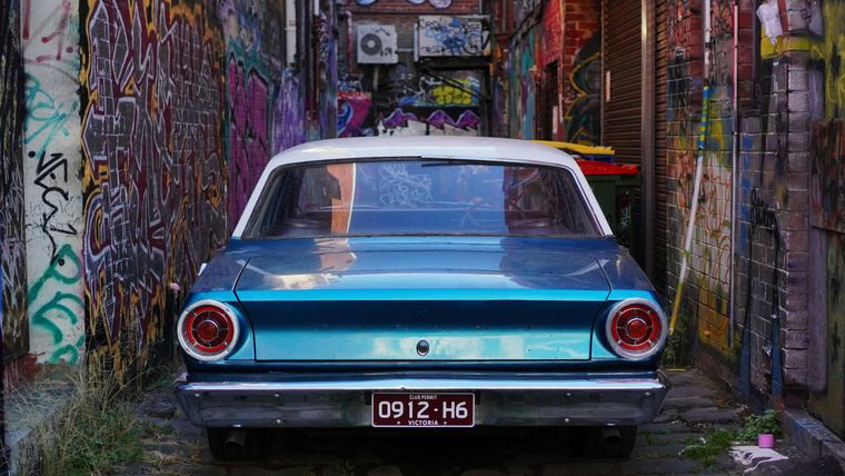 A blue 1960s Ford Falcon parked rear-on in a narrow laneway lined with graffiti, its Victorian numberplate visible below the boot.