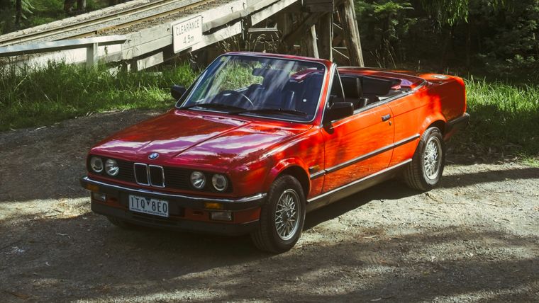 A red BMW E30 convertible parked below the Puffing Billy trestle bridge in the Dandenong Ranges as the steam train crosses above.