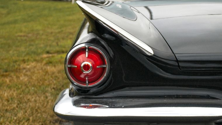 Close-up of the chrome tailfin and round red tail-light of a black classic sedan parked at a grassy kerb.