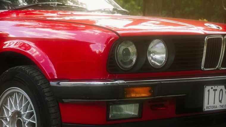 Close-up of the front of a red BMW E30, showing its twin round headlights, kidney grille and a Victorian numberplate.