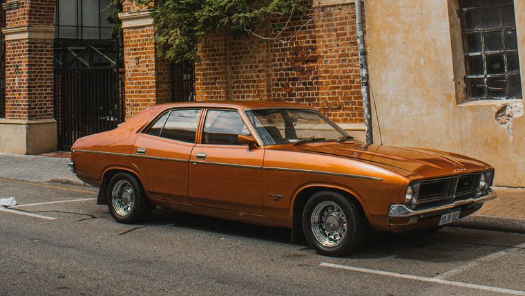 A copper Ford Falcon sedan parked at the kerb outside a heritage brick building.
