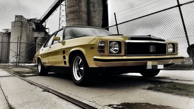 Yellow Holden GTS sedan parked in front of industrial grain silos under a moody sky.