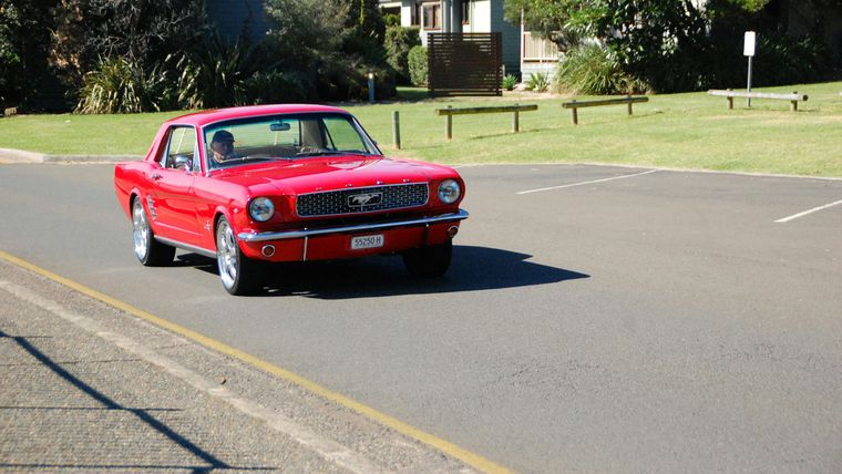 Red classic Ford Mustang coupe driving along a coastal Victorian street lined with Norfolk pines.