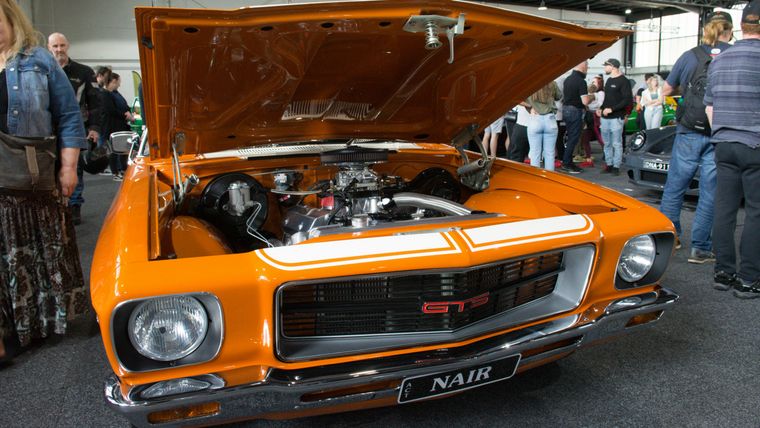 An orange Holden Monaro GTS on display at an indoor car show with its bonnet raised, showing the engine bay.
