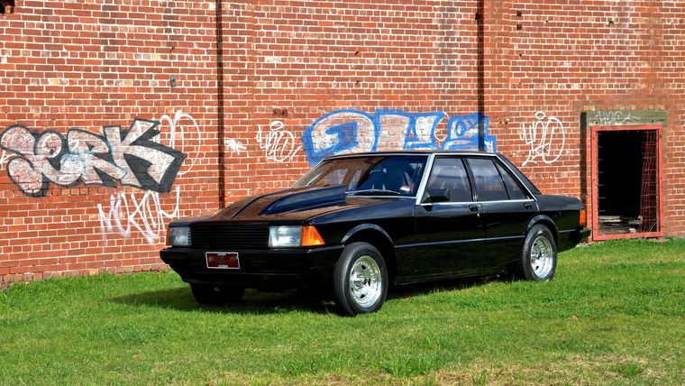 A black Ford Falcon sedan parked on grass in front of a graffitied red brick warehouse wall.