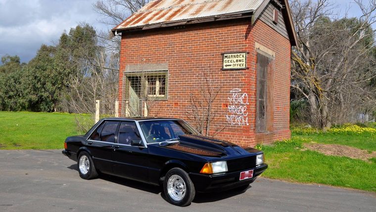 Black Ford Falcon sedan parked outside an old heritage brick building.